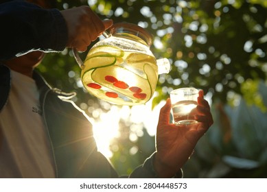 Sunlight glints through a glass pitcher. Cucumbers and red berries float inside. Hands pour liquid into a clear glass. Green leaves blur in the background. Peaceful moment, warm glow, natural setting