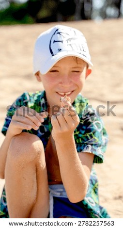 Sunlight glows on a smiling boy at the beach. He wears a white cap and colorful shirt. Hands hold a small seashell, close to his mouth. His eyes sparkle with joy and curiosity. Perfect for vacation