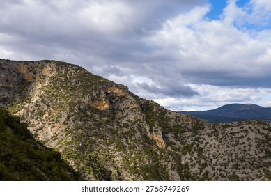 Sunlight highlights the rugged slopes and sparse vegetation of a rocky mountain ridge beneath a sky filled with dramatic clouds. The textured terrain and distant peaks create a sense of wild, untamed landscape.