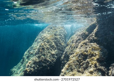 Sunlight illuminates a passage between large underwater rocks covered in algae, beneath the surface in Saint-Jean-Cap-Ferrat, France.