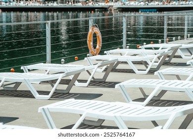 Sunlight illuminates white lounge chairs arranged near tranquil water, while a lifebuoy hangs nearby, offering safety to those enjoying the marina. - Powered by Shutterstock