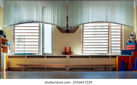 Sunlight illuminating empty gym of kindergarten through vertical blinds and wall bars, showcasing toys, basketball hoop and colorful stacked mats - Powered by Shutterstock