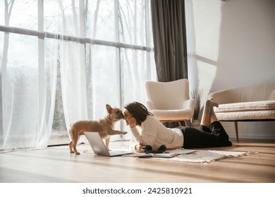 In sunlight. Lying down on floor with laptop. Looking at pet. Young woman is with her pug dog at home.