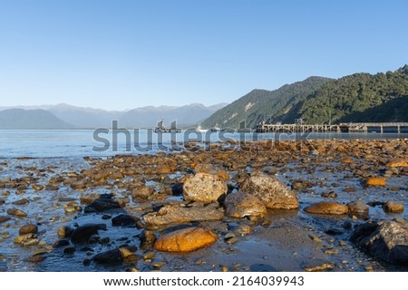 Sunlight on rocky foreshore pier with surrounding mountains as sun sets across Jackson Bay, South Island New Zealand.