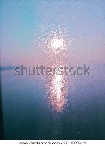 Sunlight passes through a glass surface covered with water droplets. The image shows natural light and moisture on the glass, creating a blurred view of the outside environment. No people are visible.