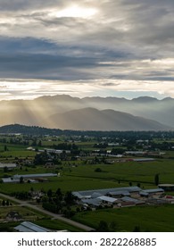 Sunlight rays pierce dramatic clouds over green farmland and misty mountains in Chilliwack, British Columbia, Canada.