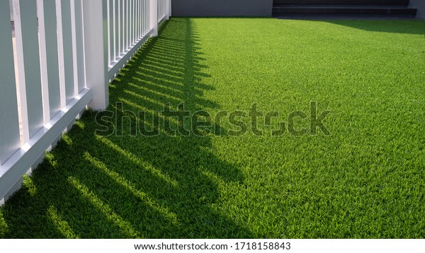 Sunlight and shadow of white wooden fence on green\
artificial turf surface in front yard of home, selective focus with\
high angle view 