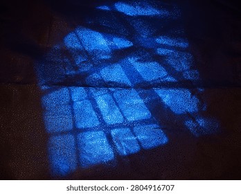 Sunlight shines through a window creating blue light patterns on the surface below. The shapes form a grid over the material. The scene shows an indoor space where light interacts with a surface.