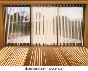 Sunlight Shining Through Full Length White Vertical Blinds In Front Of Three Glass Sliding French Doors Leading To A Patio, Garden And Sea View, Creating Shadow Stripes.