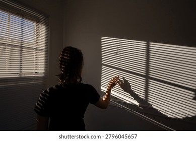 Sunlight slices through blinds, casting striped shadows. Figure stands, hand gently touching the wall. The room feels quiet, still, and warm. Light contrasts with dark, creating depth and mood. Sugges