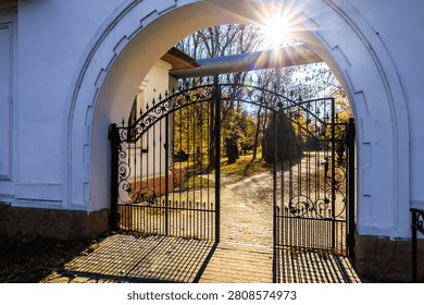 Sunlight streams through an arched gate into a quiet garden. Metalwork doors frame a winding path lined with autumn trees. Soft golden light filters through leaves, casting gentle shadows