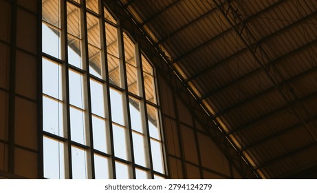 Sunlight streams through a tall, grid-patterned arched window, highlighting the textured, sloping ceiling of a historic building, creating a dramatic interplay of light and shadow.