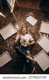 Sunlight streams through window of modern art studio on female painter artist who lying on floor near canvases at sunrise. Top view.