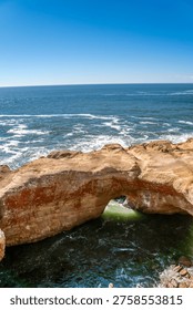 Sunlit arch frames ocean's deep blue. Weathered stone bridges sky and sea. Waves crash below with rhythmic force. Clear sky casts calm, golden glow. Ideal for travel, nature, or coastal themes