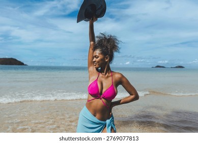 Sunlit beach, turquoise waves, distant islands. Woman in pink bikini, blue wrap, joyful pose. She holds hat high, smiling toward horizon