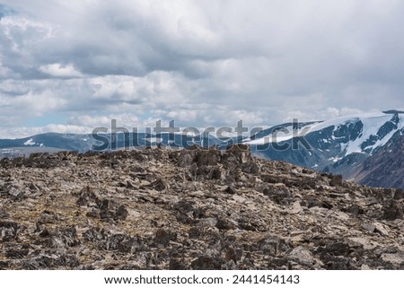 Sunlit big rocky hill of sharp stones against mountain range silhouette and snow mountains in cloudy sky. High stony pass and large snowy ridge in sunlight. Rocks on mountain top in changeable weather