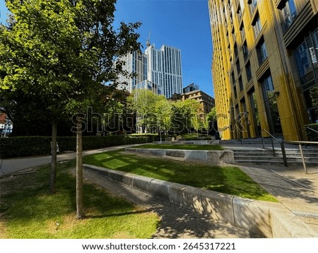 Sunlit buildings and trees create a harmonious urban green space, with modern architecture contrasting against natural greenery. The blue sky enhances the vibrant atmosphere in Leeds, Yorkshire, UK