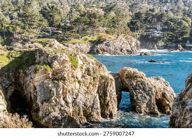 Sunlit cliffs frame turquoise ocean waves. Rugged rocks jut from water, weathered by time. Lush green trees crown distant shores. Bright sunlight illuminates every textured surface