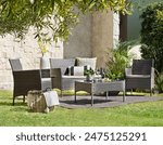 Sunlit garden dining space, a rattan weave furniture set, two armchairs and a sofa with neutral cushions, a coffee table against a textured stone wall backdrop, refreshing morning meal lush greenery.