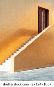 A sunlit golden wall frames a diagonal staircase leading to a dark wooden door, creating a striking architectural scene rich in texture, warmth, and subtle shadows.