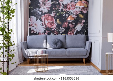 Sunlit, gray sofa by a floral print wall in the nook of a feminine living room interior with golden accessories