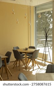 Sunlit interior dining area with table and chairs, wooden tabletop surrounded by gray upholstered chairs, large window casting sunbeams