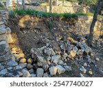 A sunlit outdoor scene showing a partially collapsed stone retaining wall with scattered rocks and soil, surrounded by trees and greenery in a terraced garden landscape.