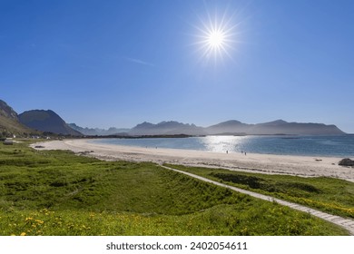 Sunlit pathway meandering through grass and wildflowers to the white sands of Rambergstranda Beach in Jusnesvika Bay, Lofoten, with a shimmering sea and mountainous horizon under a radiant sun