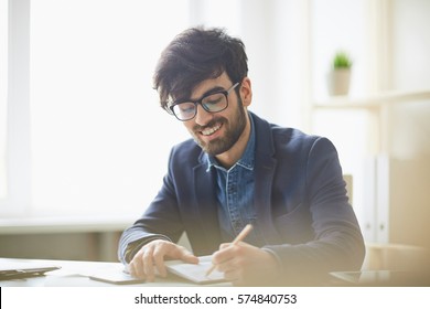 Sunlit Portrait Of Young Creative Mn In Glasses And Business Casual Wear Writing Something Down To Notebook, Smiling And Looking Pleased Sitting At Desk In Modern Office