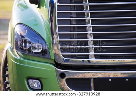Sunlit powerful modern stylish and comfortable green big rig semi truck of latest model commercial long-distance transport shiny chrome grille efficient headlight in parking lot waiting for cargo.