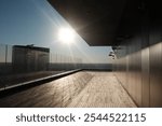 Sunlit rooftop terrace with glass railings and a wooden deck, overlooking an expansive view. The modern architecture and clear sky create a serene ambiance.