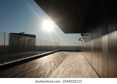 Sunlit rooftop terrace with glass railings and a wooden deck, overlooking an expansive view. The modern architecture and clear sky create a serene ambiance. - Powered by Shutterstock