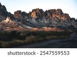 Sunlit rugged mountain range in a desert setting, featuring warm, rocky peaks and sparse vegetation under a pale, clear sky.
