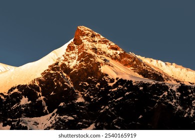 Sunlit snow-capped mountain peak under a clear, deep blue sky, displaying rugged, rocky features. - Powered by Shutterstock