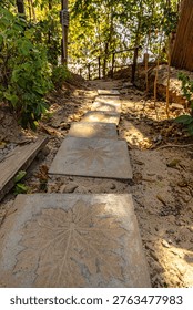  sunlit stone path of decorative leaf-pattern stepping stones winds through a shaded tropical forest or garden, creating a peaceful, rustic walkway leading toward a secluded beach. Tropical trail