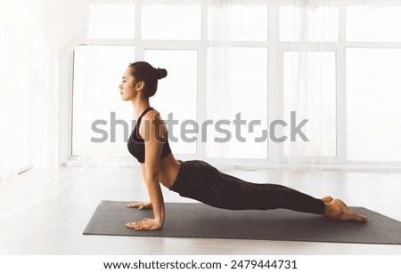 In a sunlit studio with large windows, lady is engaged in a yoga practice, focusing on an upward-facing dog pose, a common back-bending exercise in various traditions of yoga