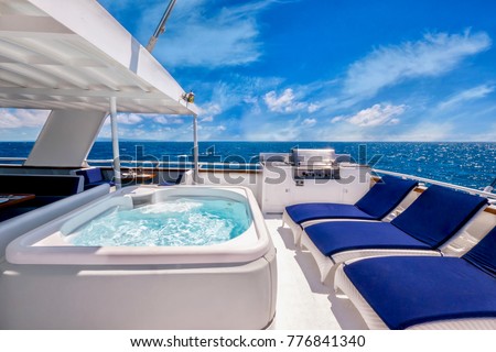 Sunny aft deck of a large, luxurious private motor yacht featuring a hot tub spa, deck chairs, and a barbecue. Blue skies and tropical water add to the concept of living the good life.