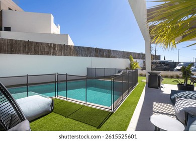 A sunny backyard featuring a rectangular swimming pool with a child safety fence, artificial grass, and cozy outdoor furniture.