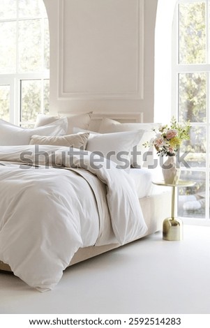 Sunny bedroom with two large windows. Four pillows, cushion and a fluffy down comforter on a double bed with bedside table and bouquet in vase on it. Side view close-up photo. Suitable for mockup