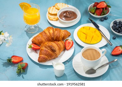 Sunny continental breakfast food on blue concrete table, with beautiful tropical plants shadows, coffee, croissants, orange juice, berries, mango, morning time holiday vacation concept