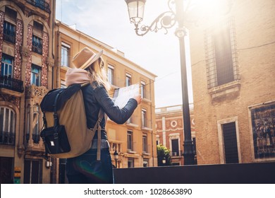 Sunny day. Back view. A young tourist woman in a hat and with a backpack is standing on a city street and is looking for a way on the map. A girl traveler walks the city, looks at the sights.