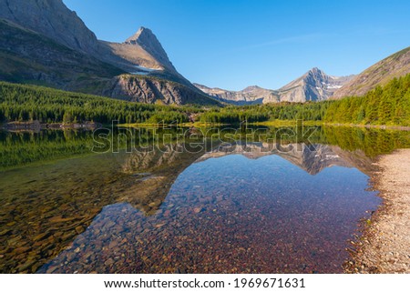Sunny day by Bullhead lake in the Glacier National Park. Mountain range reflection in water surface. Red pebbles on the beach. Hiking to Swiftcurrent Pass in early, sunny morning in American Rockies.