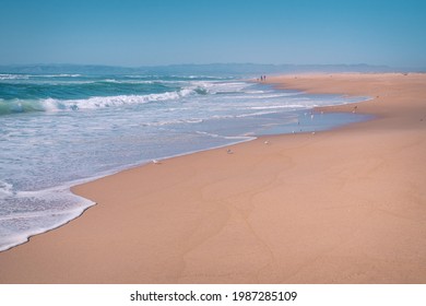 Sunny Day On The Beach, Beautiful Seascape Background. Turquoise Colored Sea, Wide Sandy Beach, Silhouette Of Walking People On A Horizon, And Clear Blue Sky