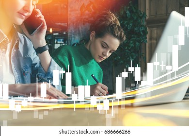 Sunny day. Two young hipster businesswomen sitting in office at table and working together.On table laptop. First woman signs documents,second talking on phone. On foreground binary options charts.