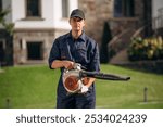 At sunny daytime, using leaf blower. Professional gardener is on the lawn.