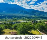 A sunny elevated view of a river valley with distant village rooftops, dense green forest, and steep blue mountain ridges under a partly cloudy sky.
