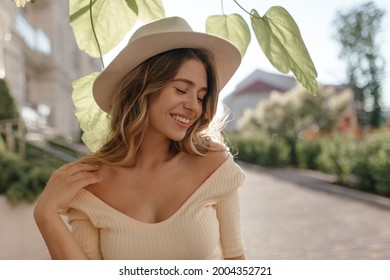 Sonniges Bild einer jungen stylischen Frau, die auf der Straße steht, in einem modischen Hut, Nahaufnahme. Sie hat sanftes Lächeln und geschlossene Augen. Schöne Halskette und nackte Schultern.