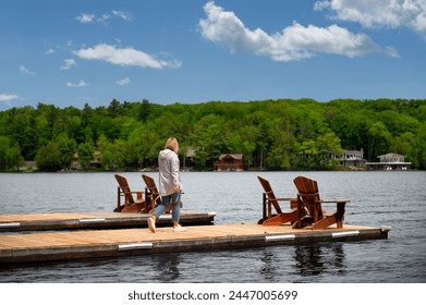 Sunny Muskoka Morning: A young woman walks toward two Adirondack chairs on a wooden dock, overlooking the serene lake. Rustic cottages nestled among trees complete the tranquil scene. - Powered by Shutterstock