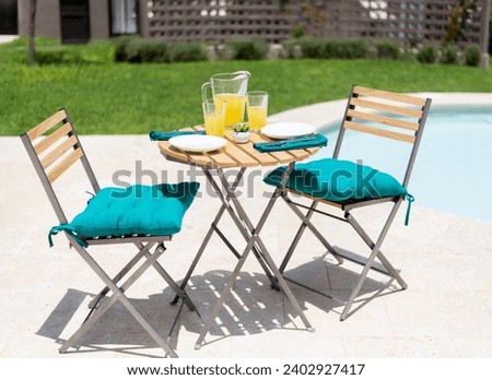 Sunny Poolside Dining Setup with a Bistro wooden Table, Teak and Metal Folding Chairs with Plush Cushions, Complete with a Refreshing Pitcher of Orange Juice and Casual Diningware on a Limestone Patio