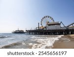 Sunny Santa Monica Pier with Ferris wheel by the ocean under a clear blue sky, waves lapping the shore.